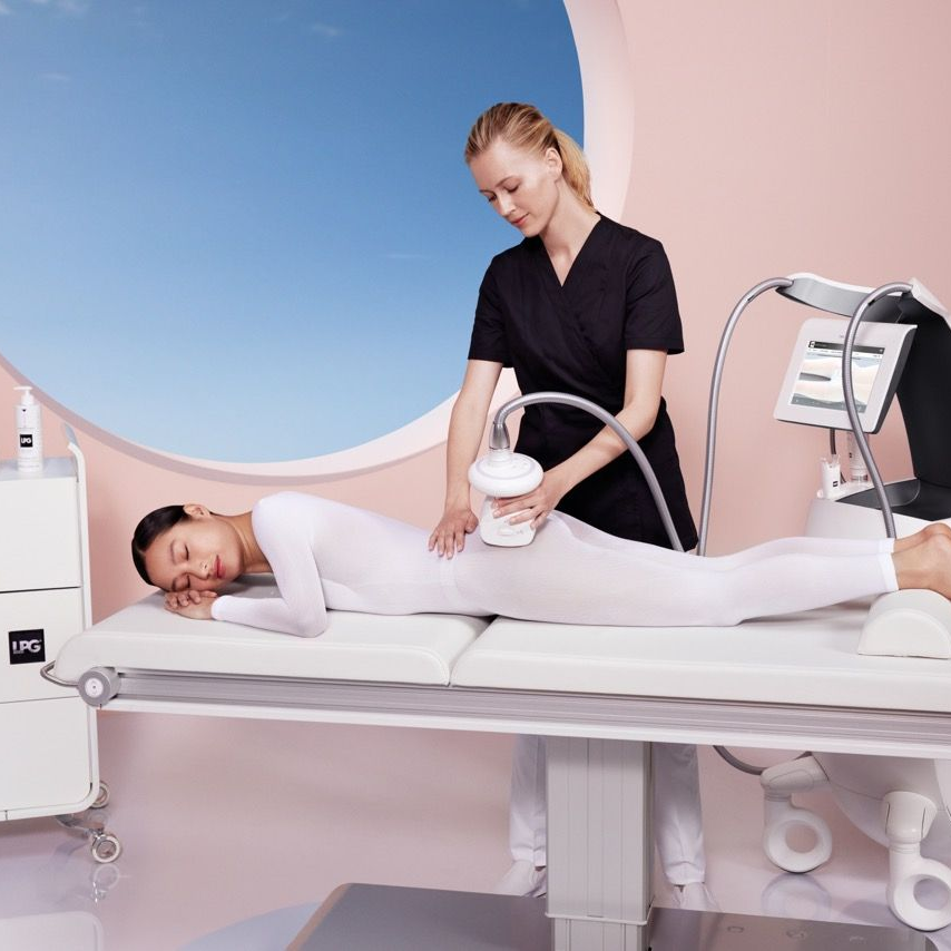 Femme recevant un soin corporel sur une table de spa ; technicienne utilisant l'appareil. Chambre rose clair, vue sur le ciel bleu.
