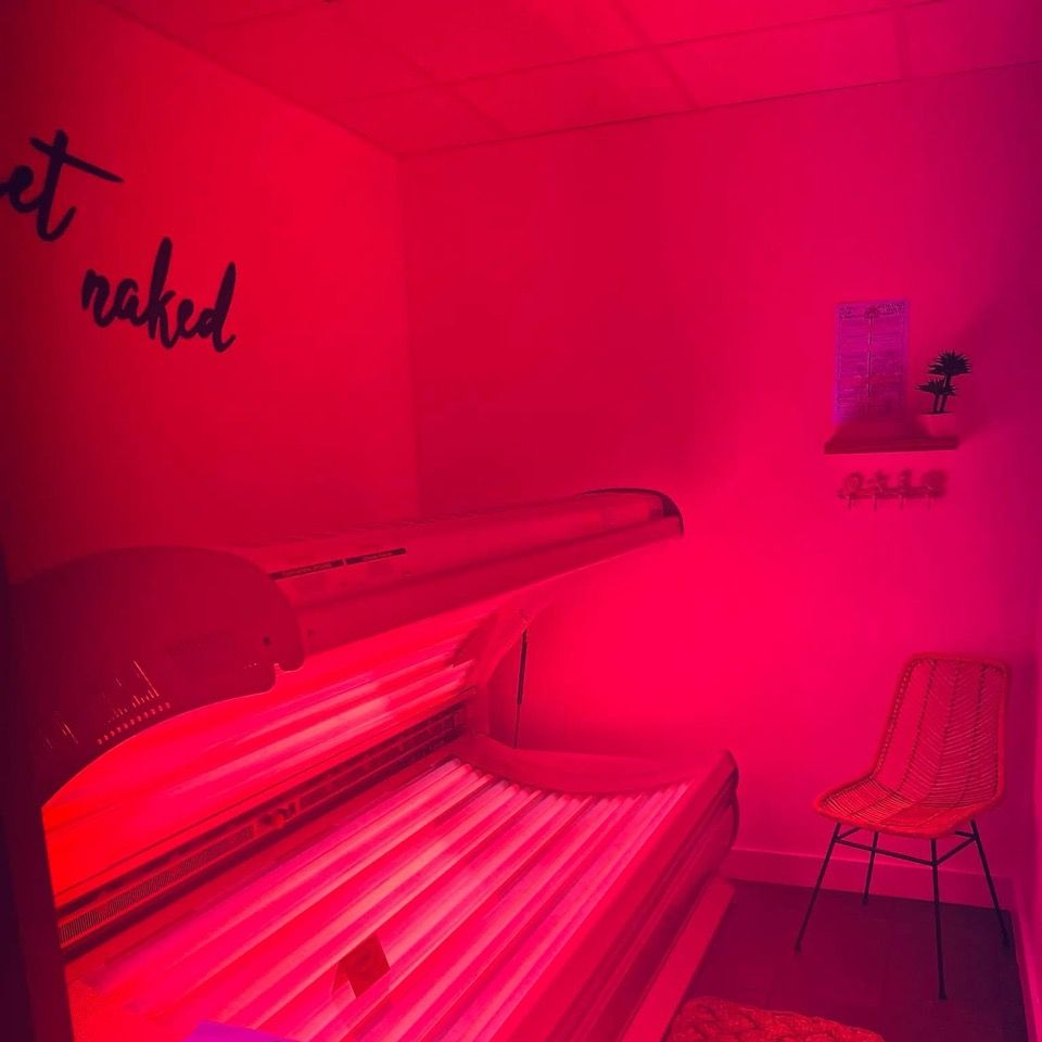 Un solarium dans une pièce éclairée en rose, avec un panneau « Déshabillez-vous ». Une chaise et une étagère avec une plante complètent le décor.