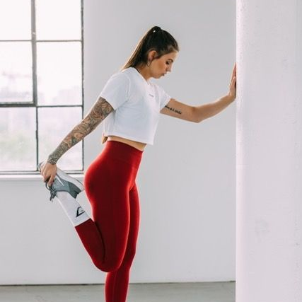 Une femme en leggings rouges étire sa jambe contre un pilier blanc ; elle porte un haut court blanc.