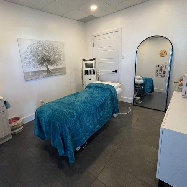 Salle de soins de l'esthéticienne : murs blancs, couverture bleu sarcelle sur le lit, grand miroir, œuvres d'art, sol gris.