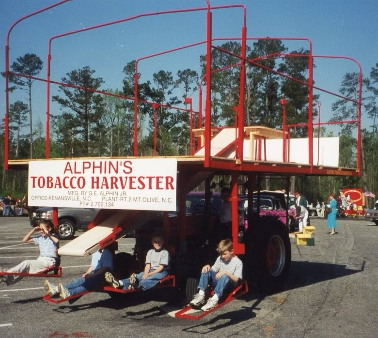 Alphin's Tobacco Harvester with children sitting on platforms. Red and white structure, bright sunny day, outdoors.