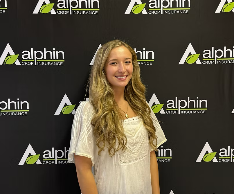 Young woman with long blonde hair and a white top smiles in front of a backdrop with the 