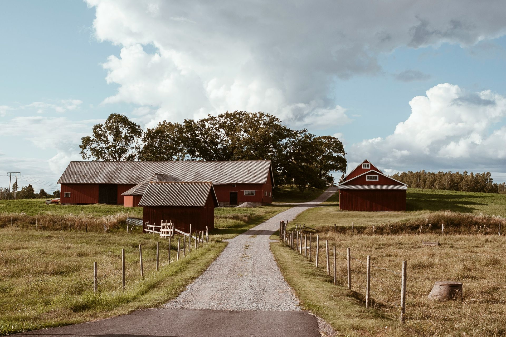 Red farm buildings with gray roofs line a gravel driveway, under a cloudy blue sky.