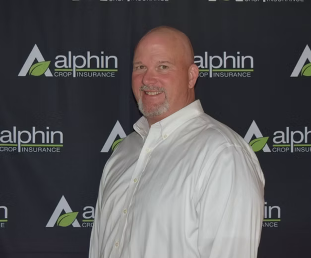 Man with a shaved head and beard in a white shirt smiles in front of an Alphrin Crop Insurance backdrop.