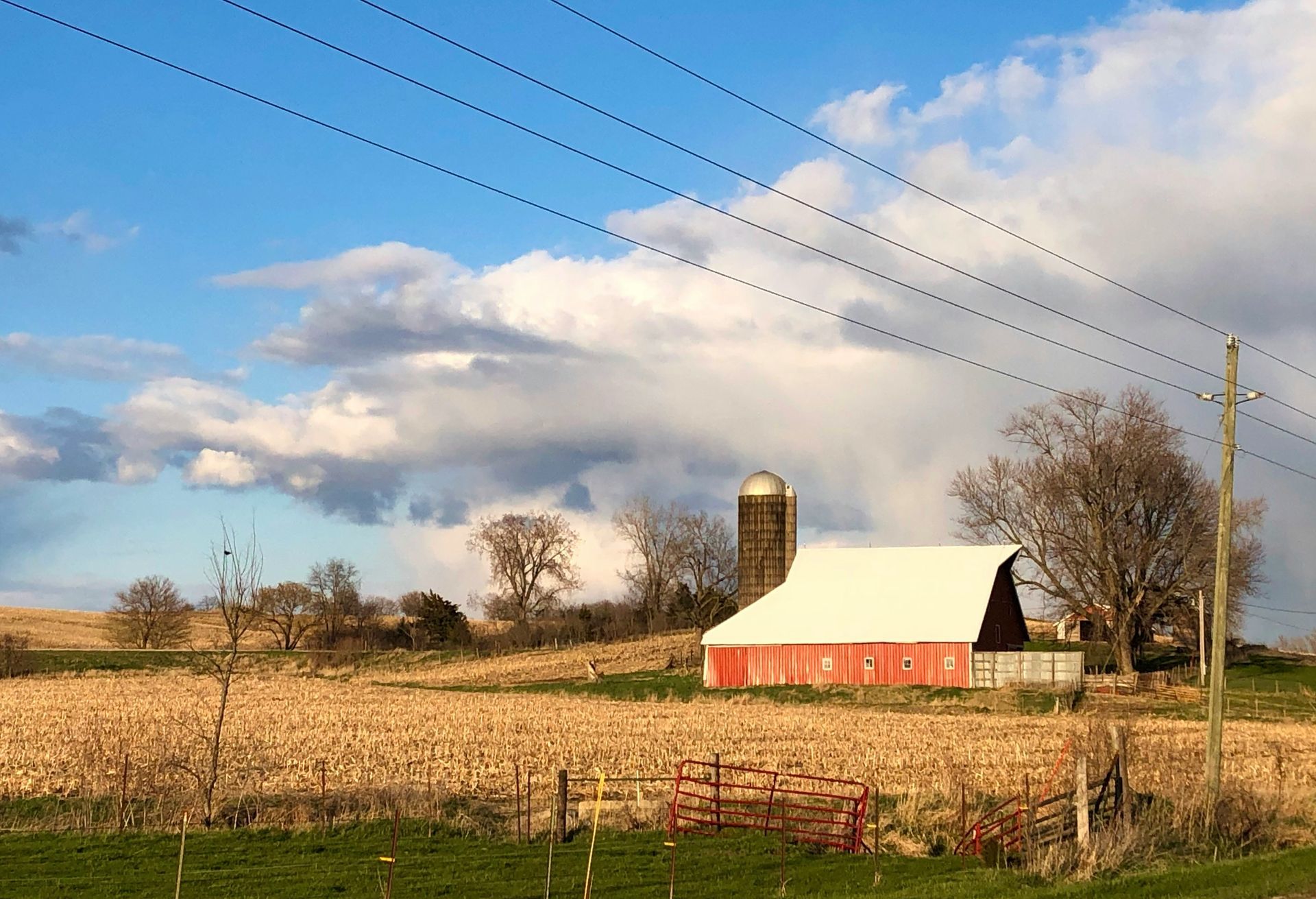 Red barn with a white roof, silo, and bare trees in a harvested field under a cloudy blue sky.