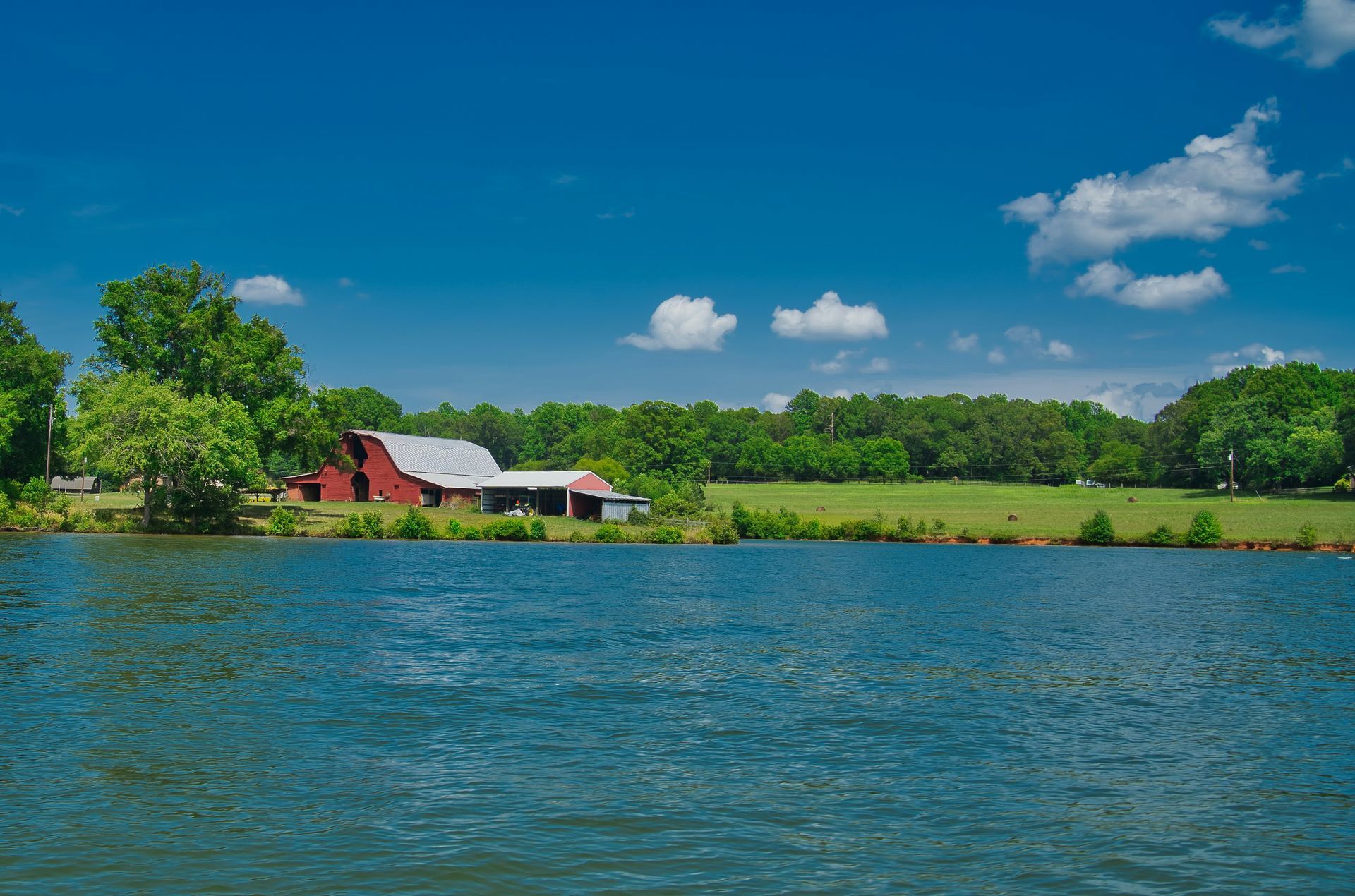 Red barn and shed beside a lake, under a blue sky with fluffy white clouds; trees and green grass surround the scene.