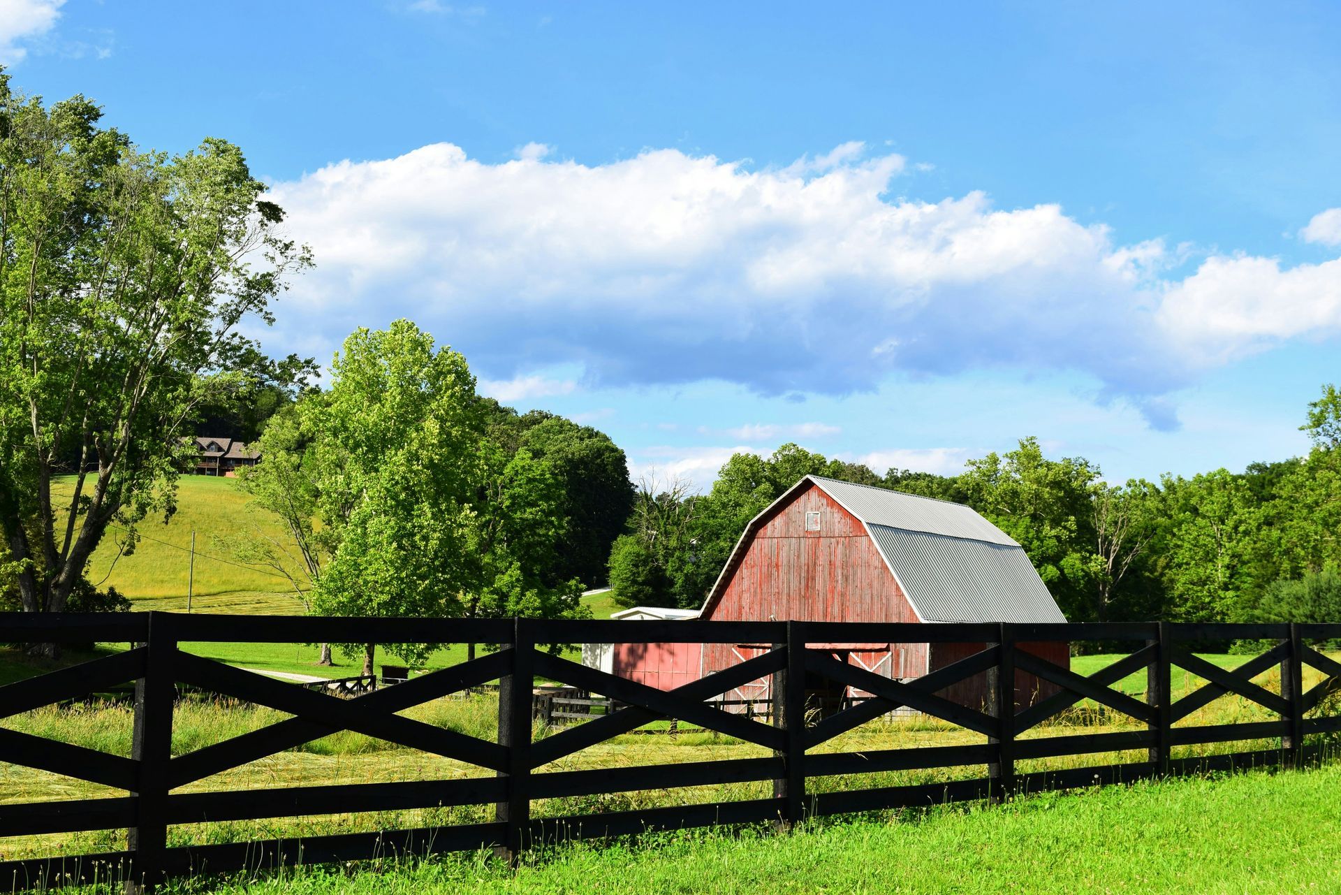 Red barn behind a black fence in a green field, under a blue sky with puffy white clouds.