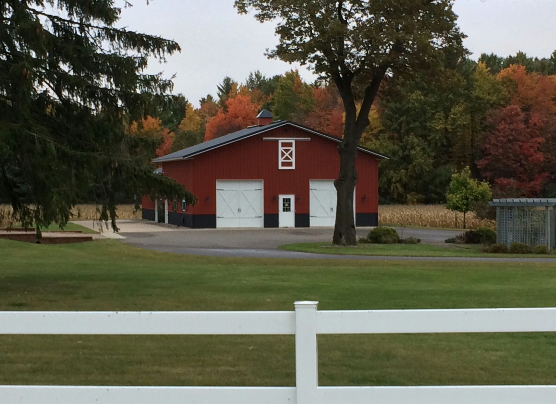Red barn with white doors and a decorative top, set against autumn foliage and a white fence.