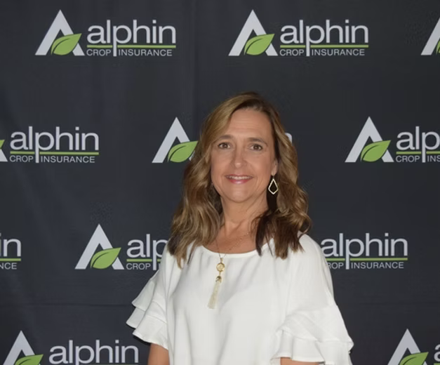 Woman with brown hair smiles in front of a backdrop with the Alphindrop Insurance logo. She wears a white shirt.