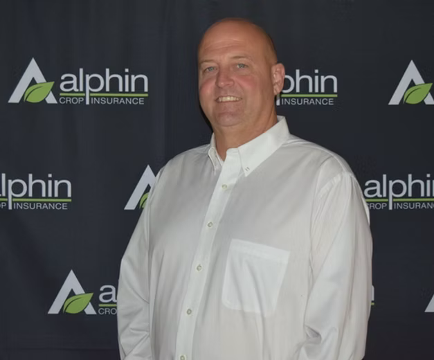 A man in a white button-down shirt smiles in front of a backdrop with the 