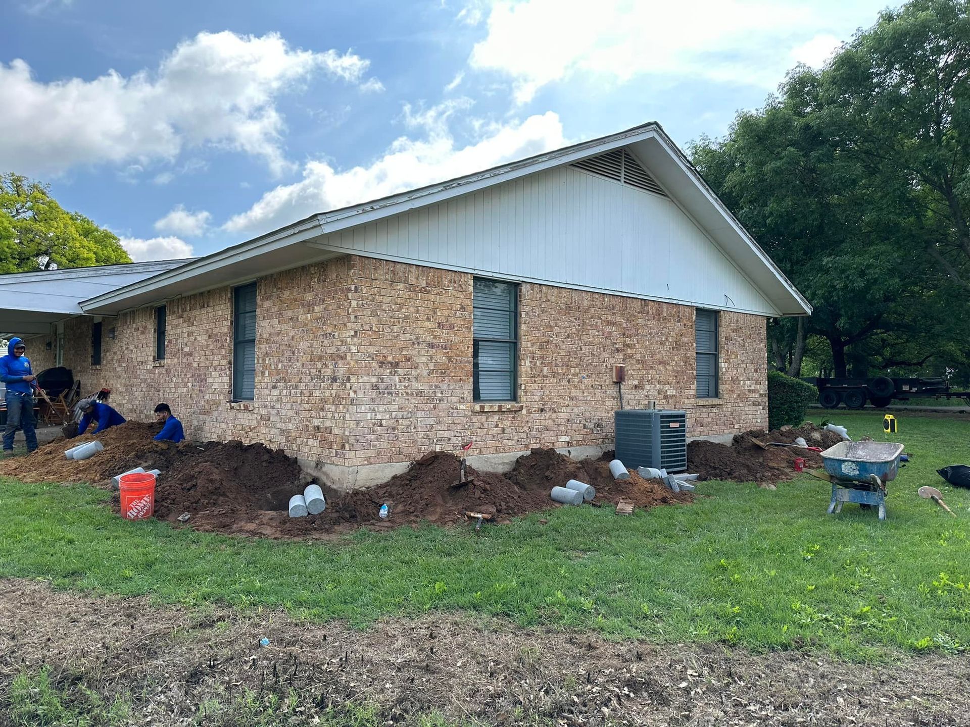 People work in a yard next to a brick house with white siding and a metal roof while landscaping.