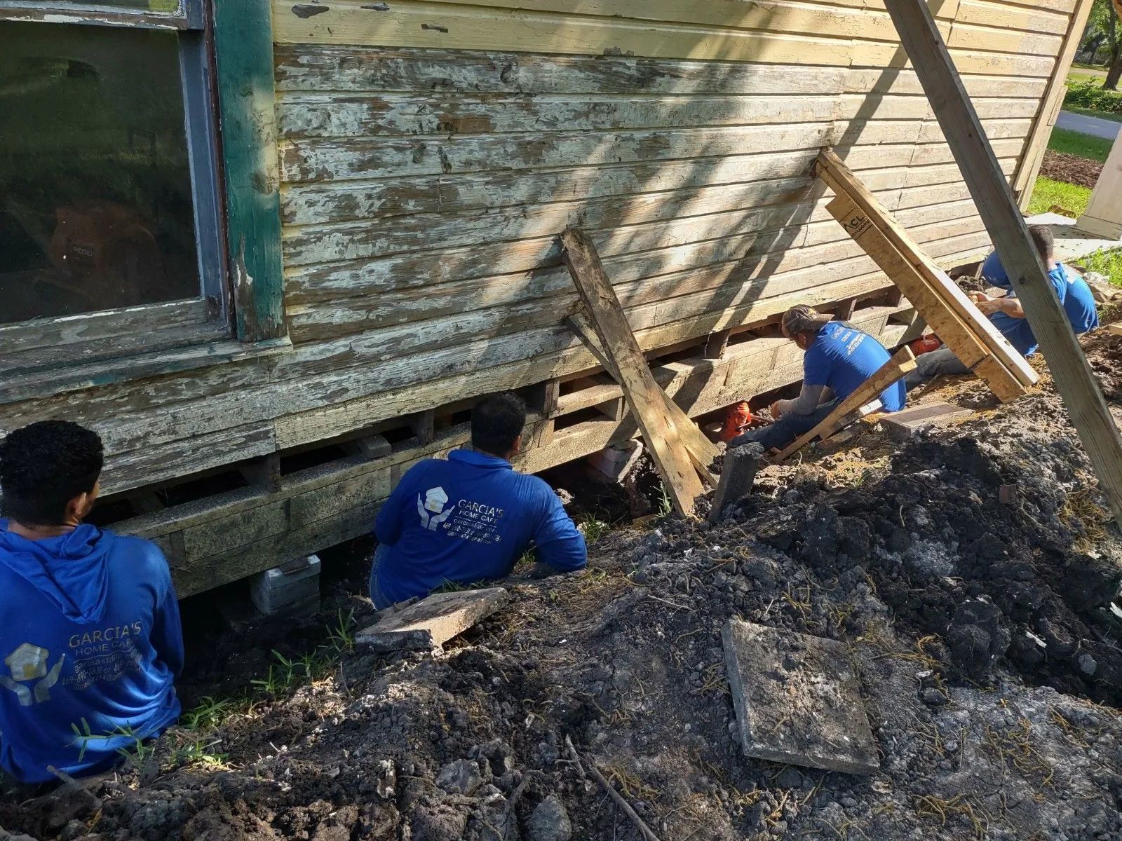 Workers in blue shirts under a house with damaged siding, supported by wooden beams, digging in dirt.