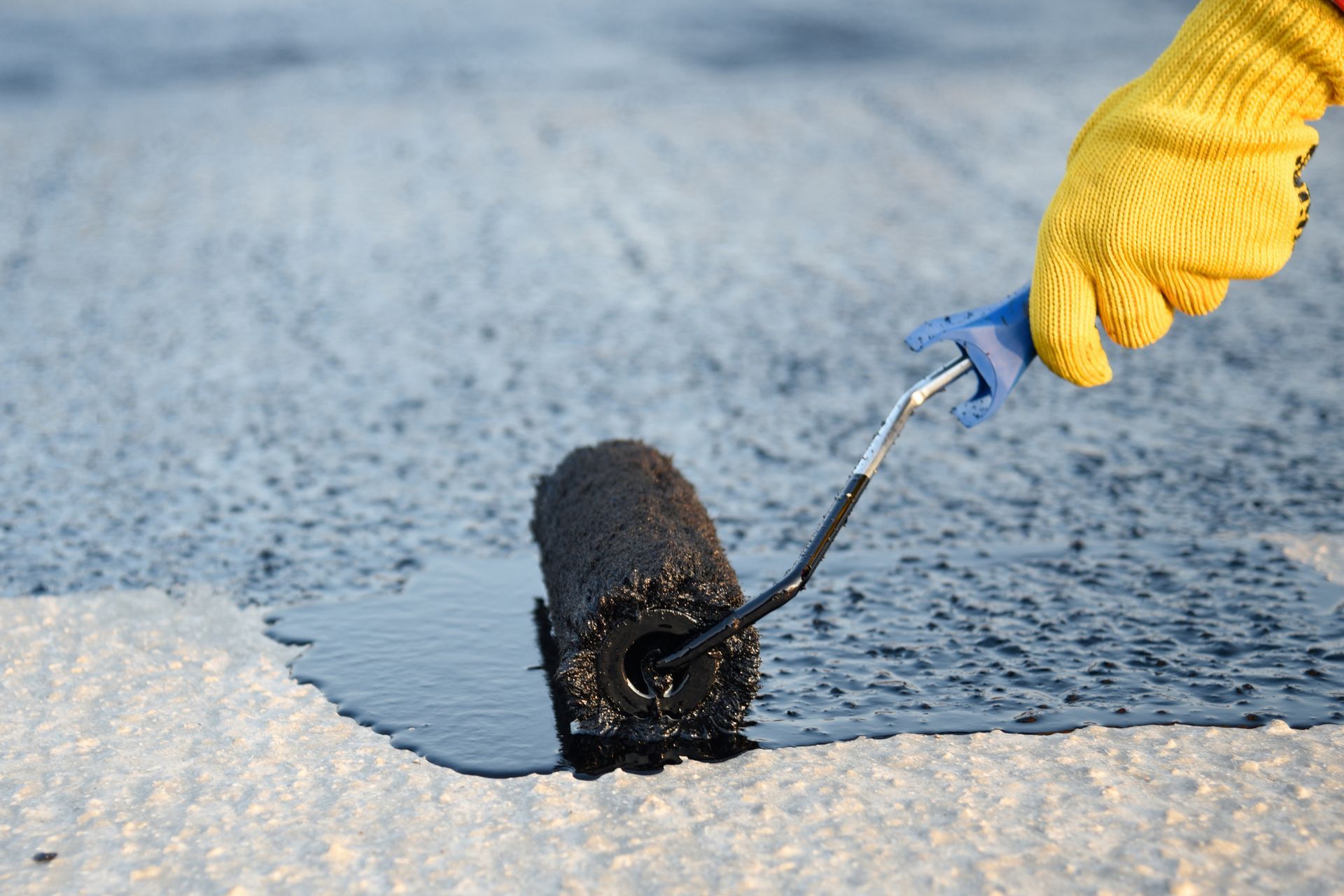 Hand in yellow glove using a paint roller to apply black sealant to a light-colored surface.