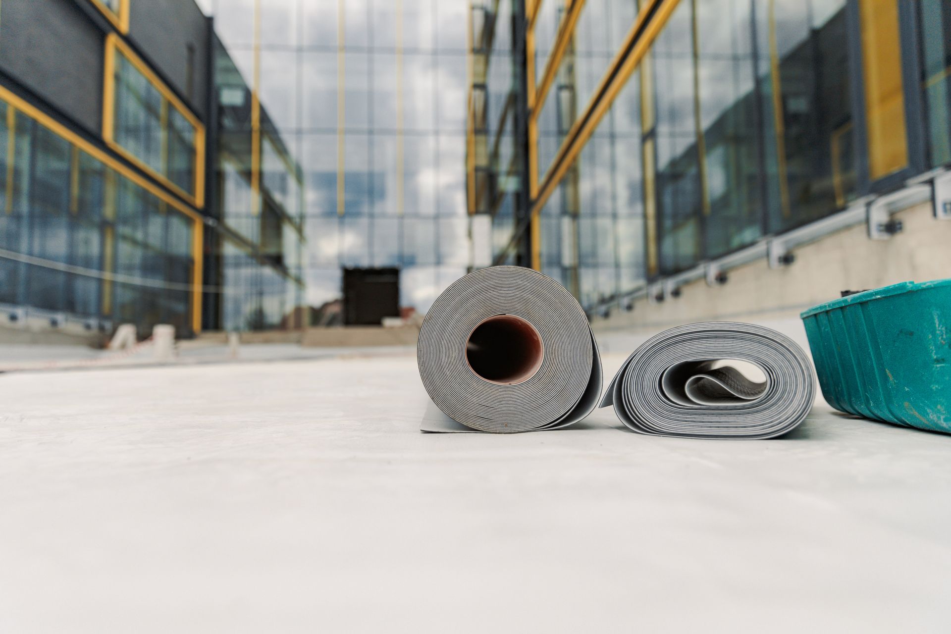 Two rolled-up gray roofing materials on a concrete surface. A modern building with reflective windows is in the background.