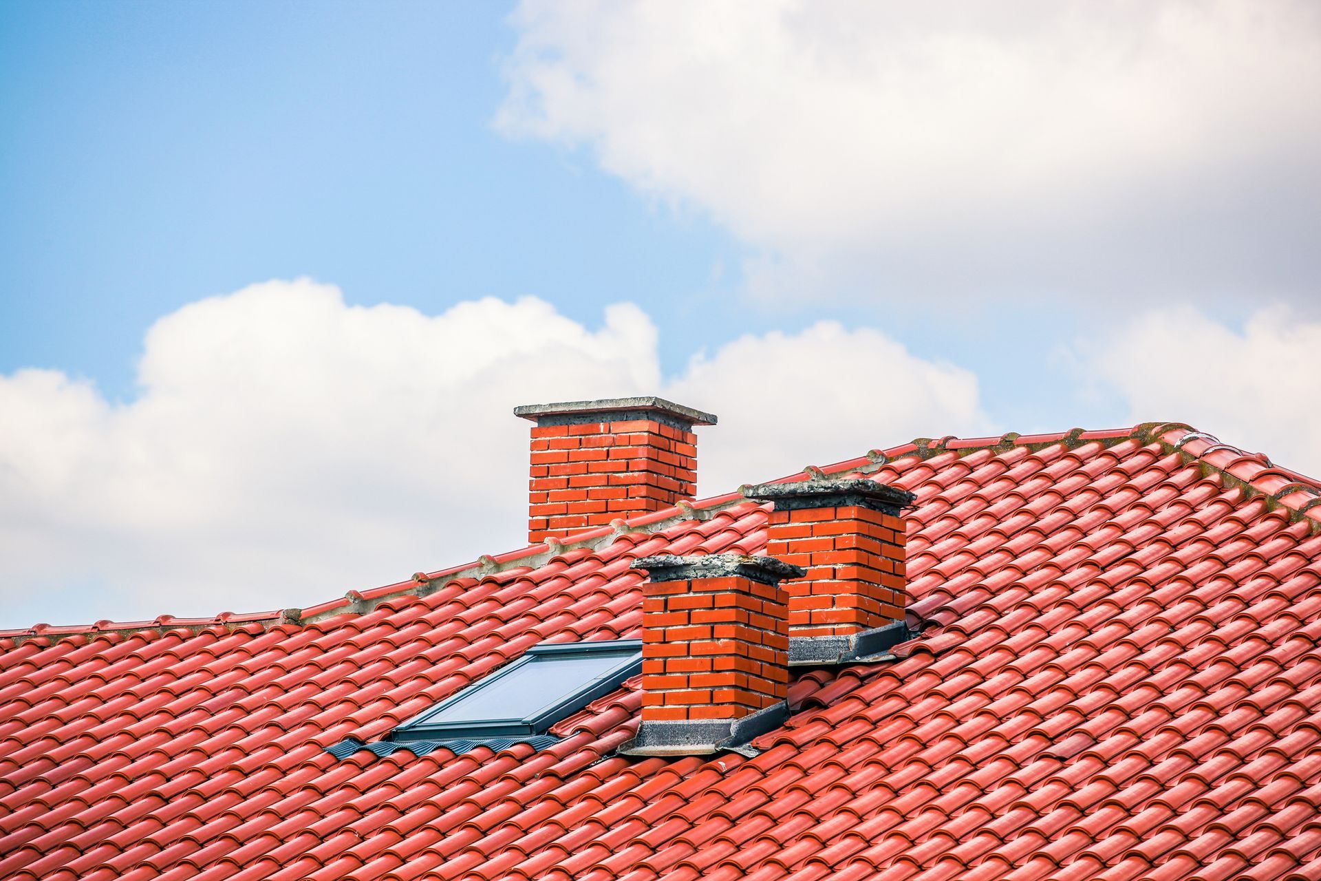 Red tile roof with three brick chimneys and a skylight against a blue sky with fluffy white clouds.