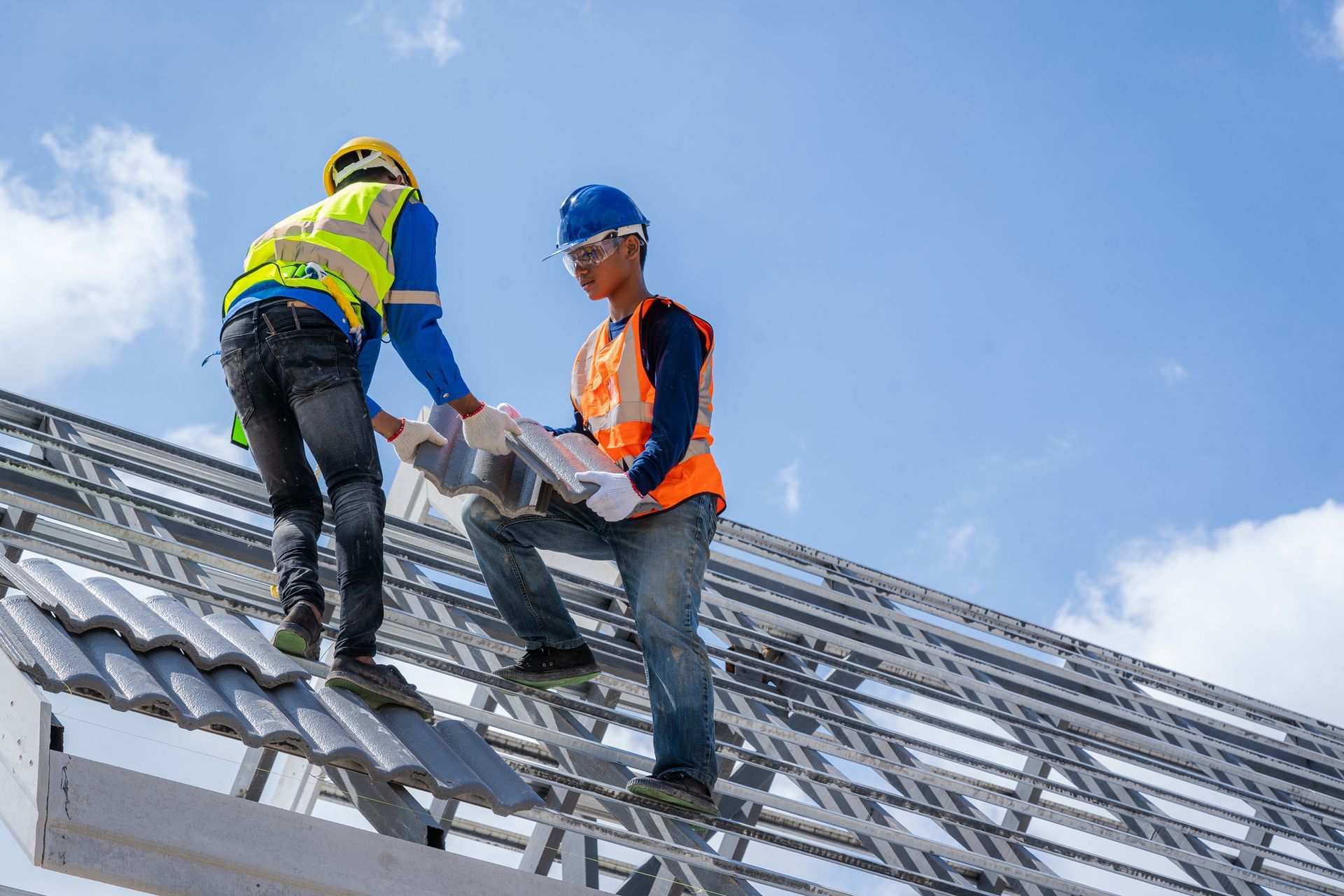 Two construction workers on a rooftop, installing roofing tiles. They wear safety vests, hard hats, and are working against a blue sky.