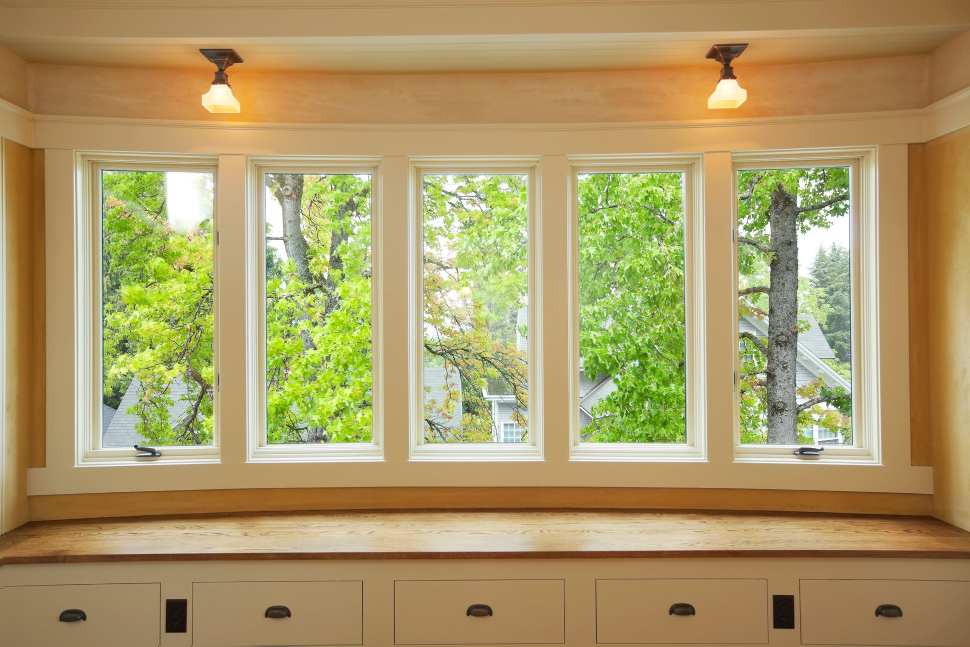 Five white-framed windows look out onto green trees and a house; a wooden bench and white drawers sit below, with two small lights above.