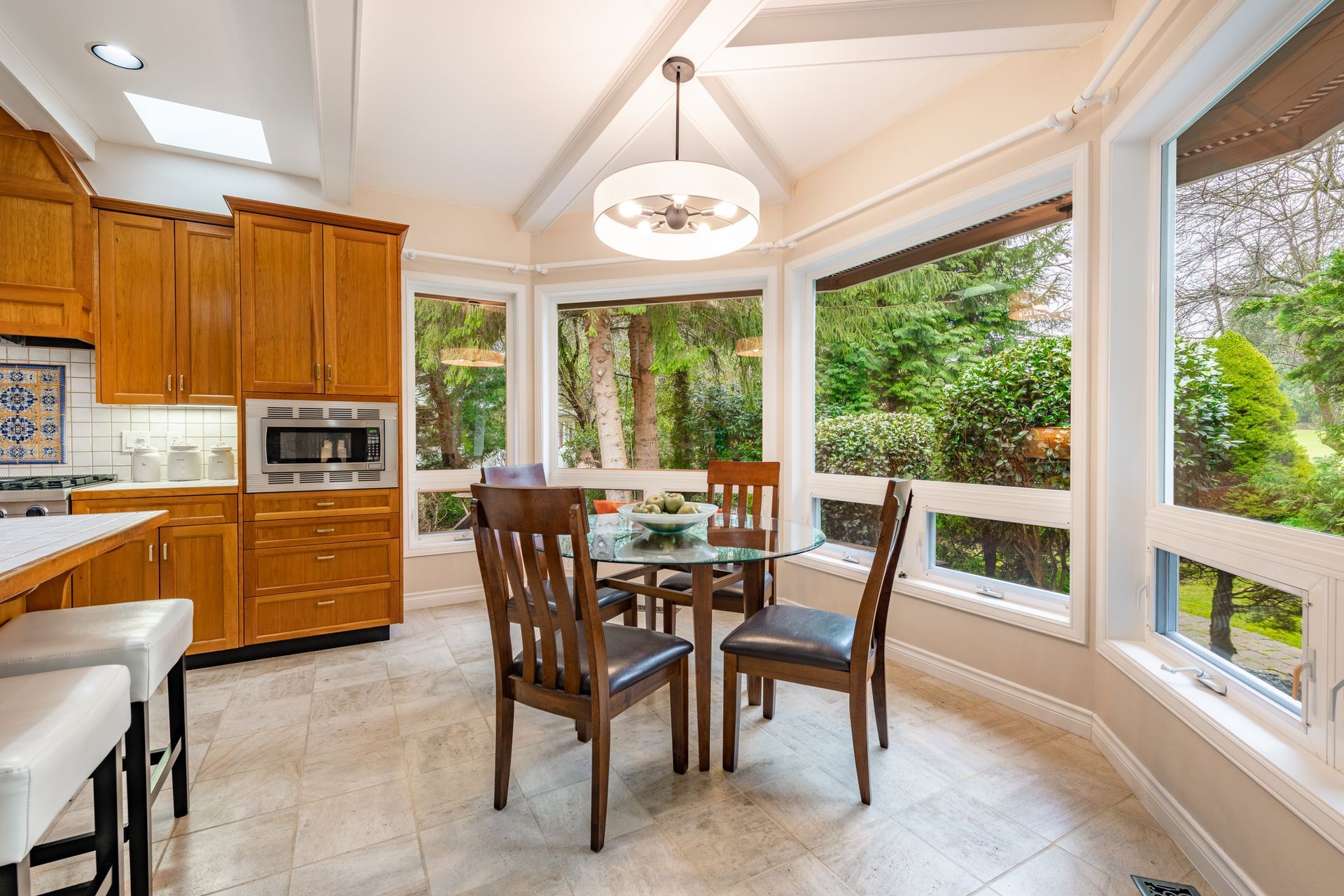 A bright kitchen with a small dining area, oak cabinets, a window view of greenery, and beige tile flooring.