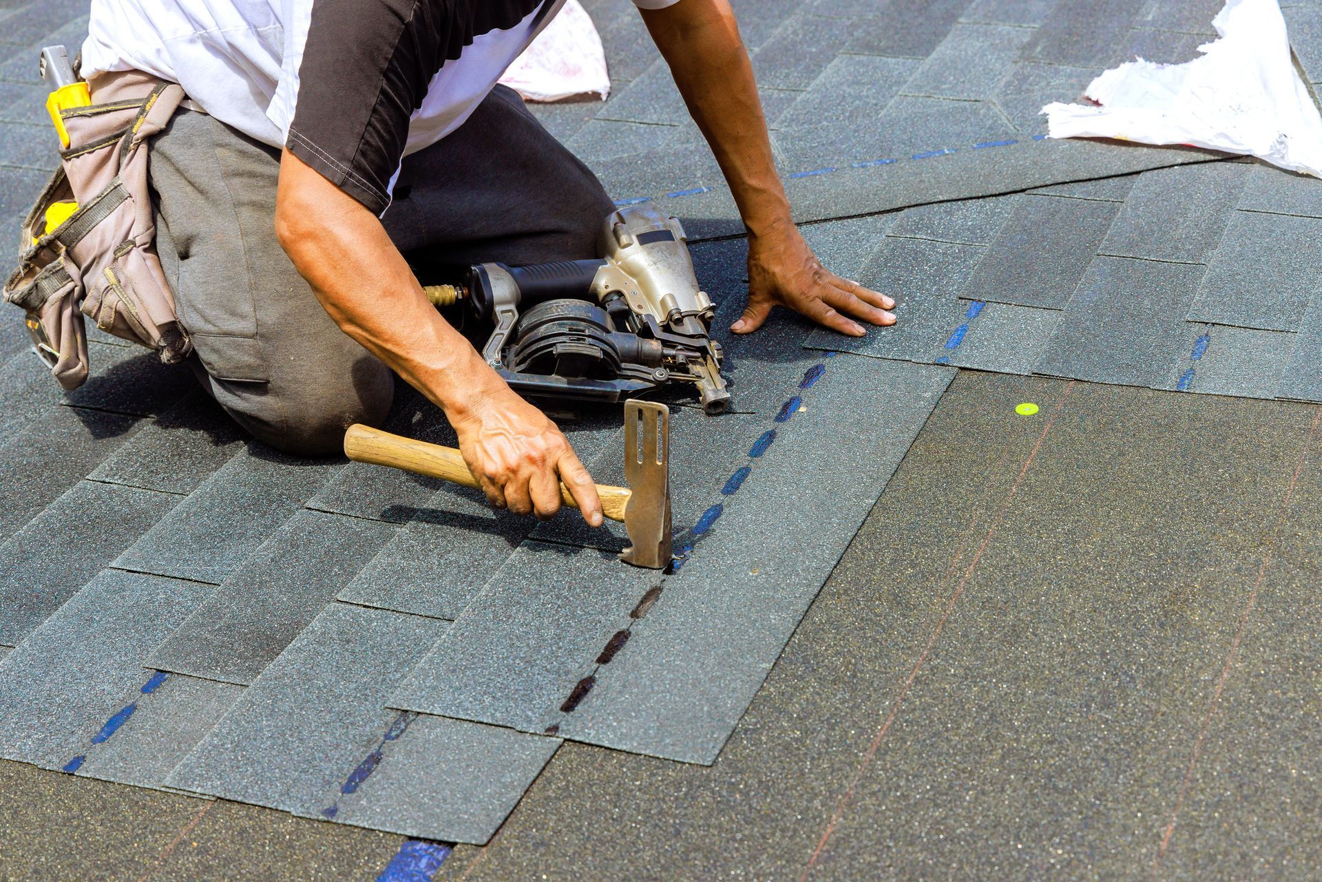 Roofer kneeling on a dark shingled roof, hammering a roofing shingle in place. He is wearing work clothes and safety gear.