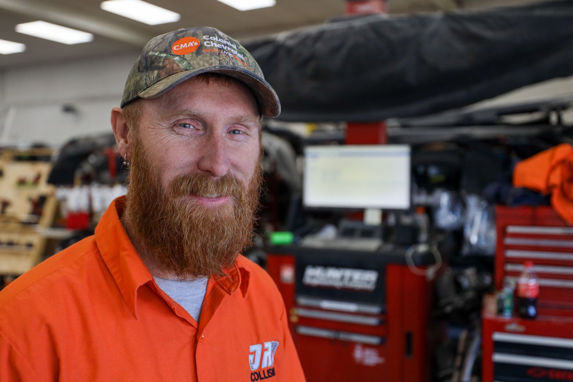 A man with a beard wearing an orange shirt and hat is standing in a garage.