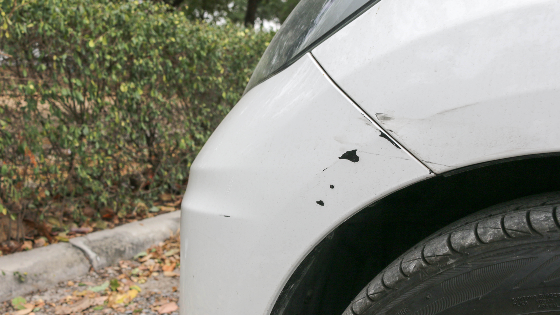 A white car with a damaged bumper is parked on the side of the road.