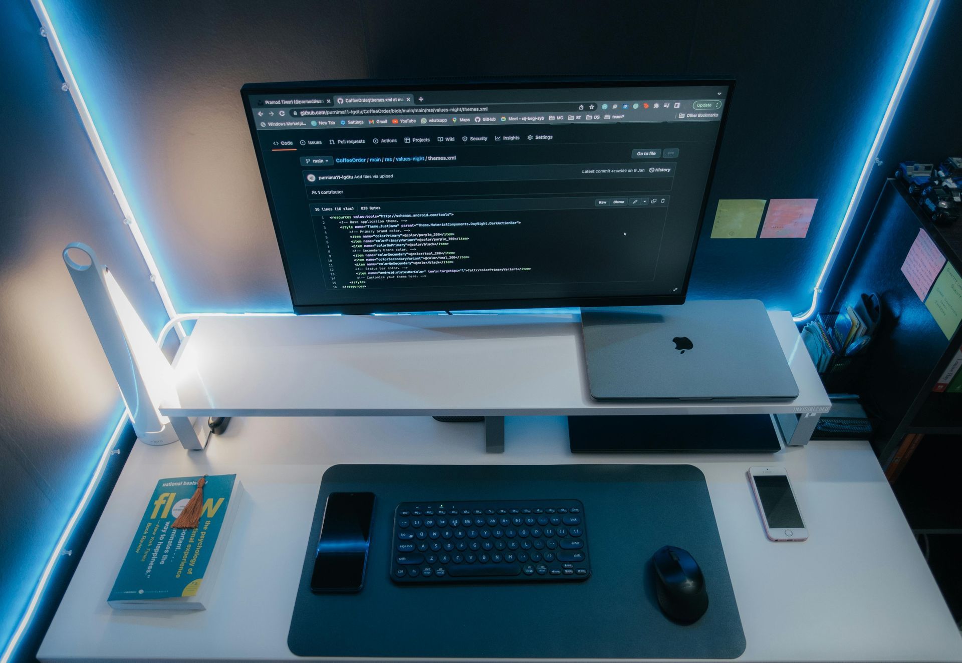 A modern desk setup with a monitor displaying code, a laptop, keyboard, and phone, illuminated by blue LED lights.