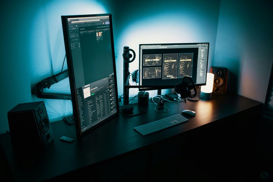 A dark workspace with dual monitors, a keyboard, and speakers under blue lighting.