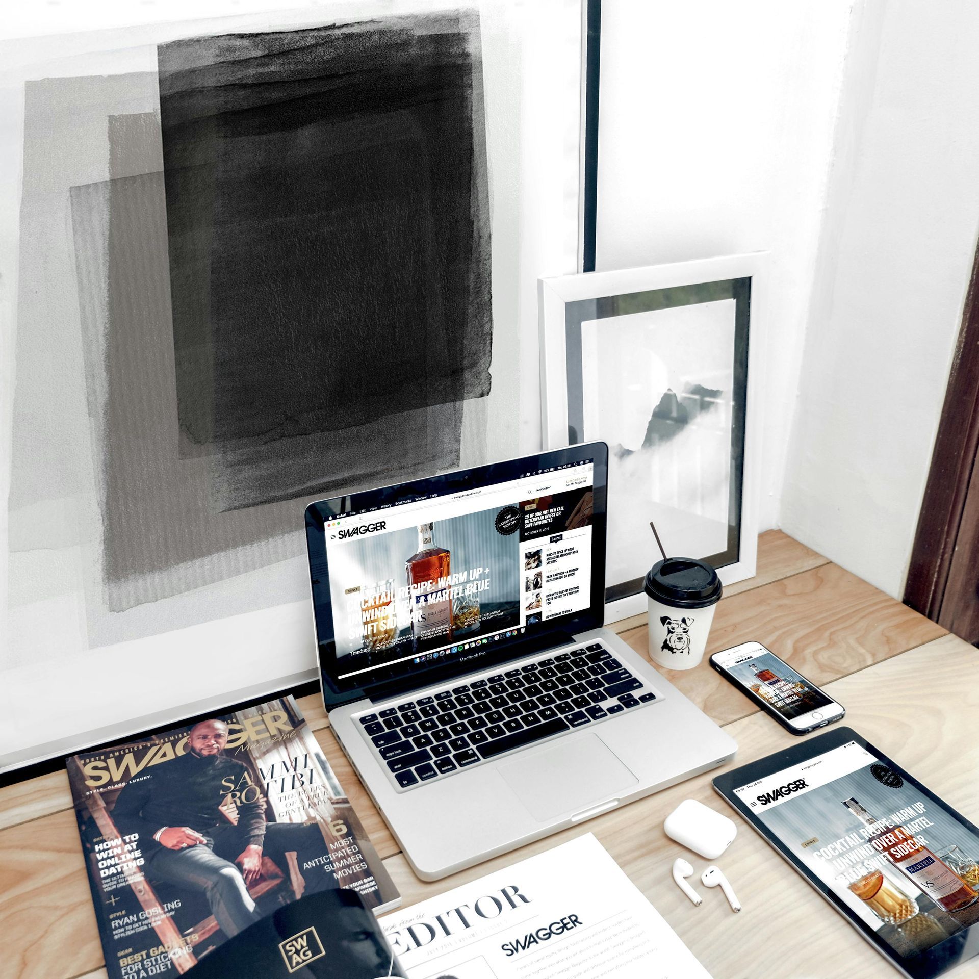 Laptop and other devices on a desk, with magazines, a coffee cup, and modern art on the wall.