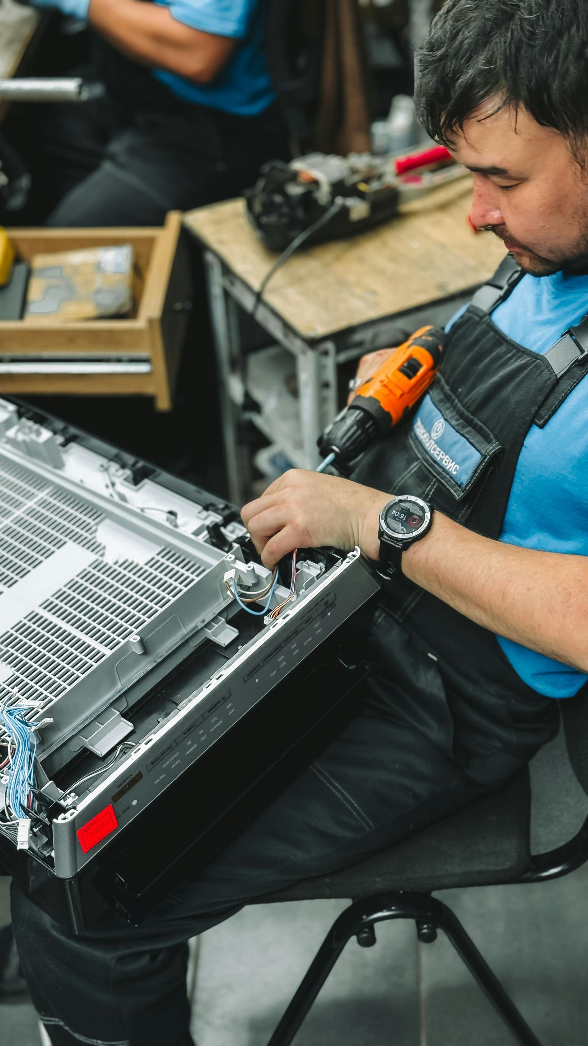 Man in blue shirt and overalls assembling a device with an orange drill, indoors.