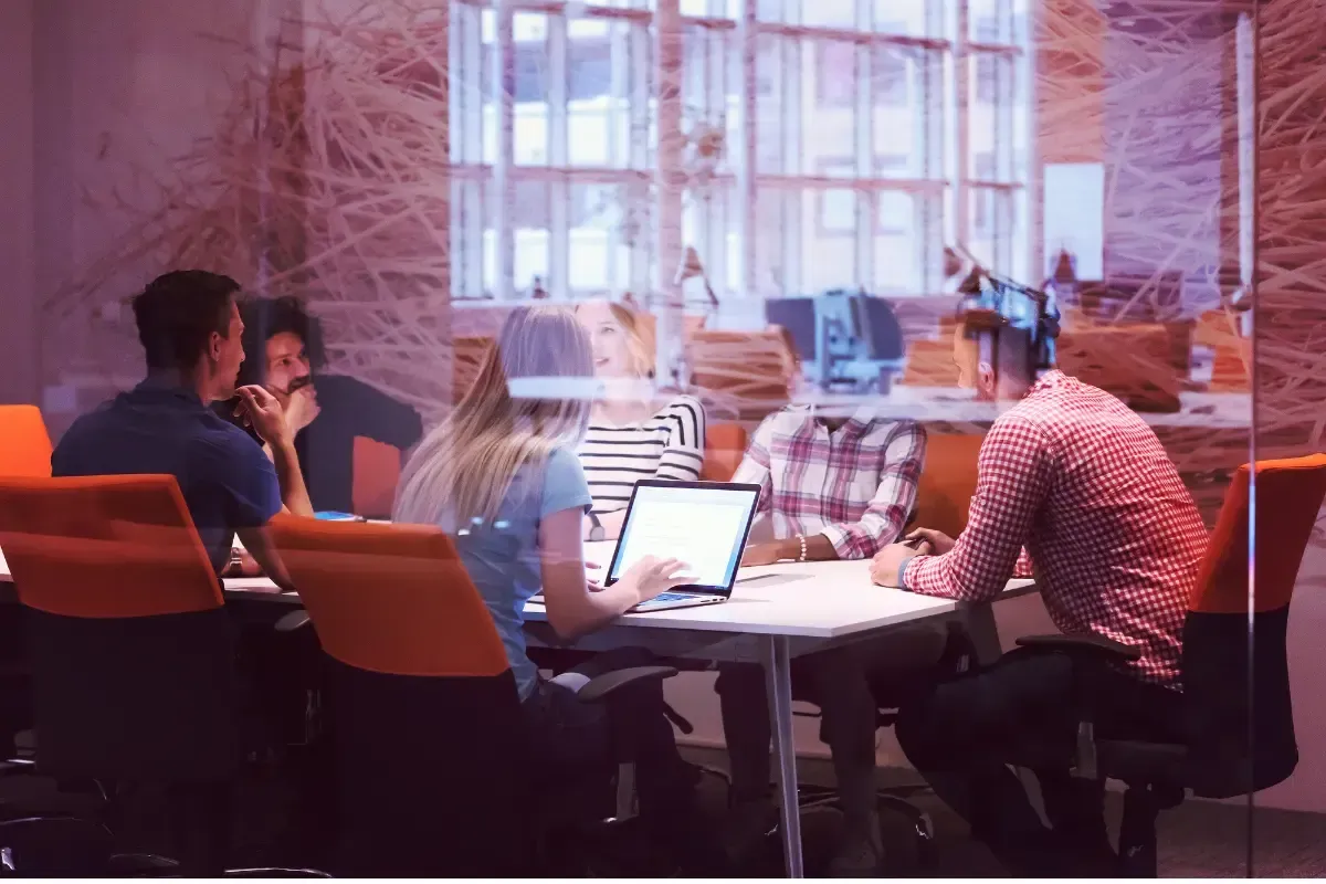 People at a table in a modern office, laptop open, discussing. Glass wall, orange chairs, bright light.