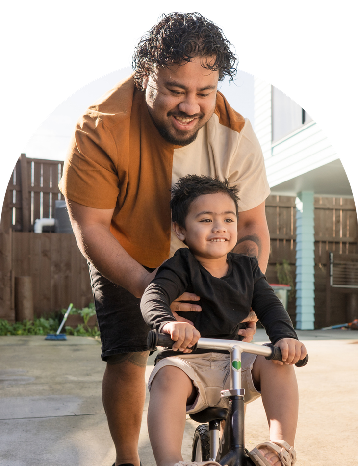 A man helps a young child ride a small bicycle. The child smiles as the man guides from behind, in a sunny outdoor setting.