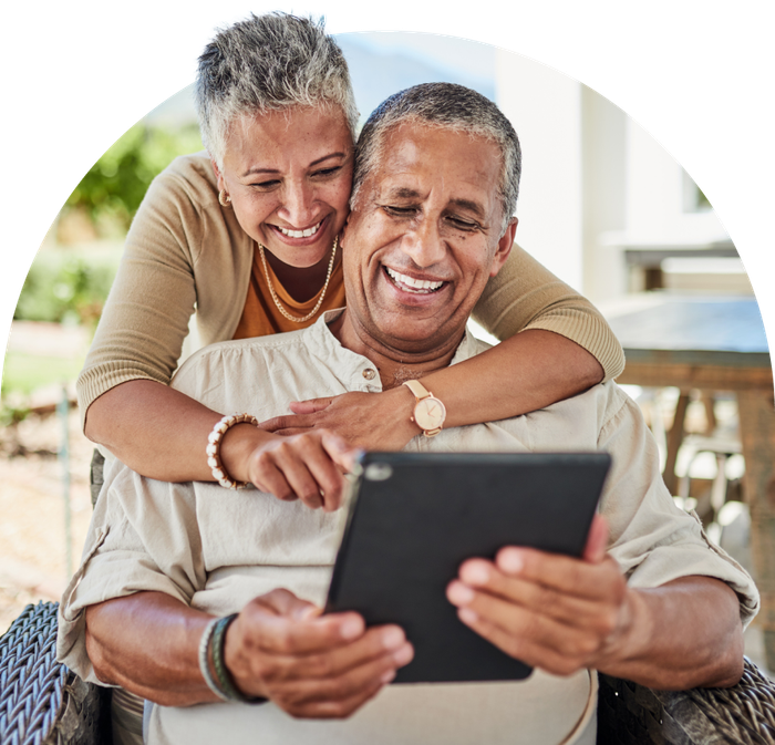 Smiling older couple looking at a tablet together outdoors. Woman has her arms around the man, who is holding the device.
