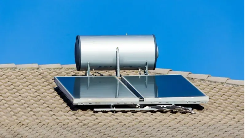 A silver solar water heating system mounted on a shingled roof against a clear blue sky.