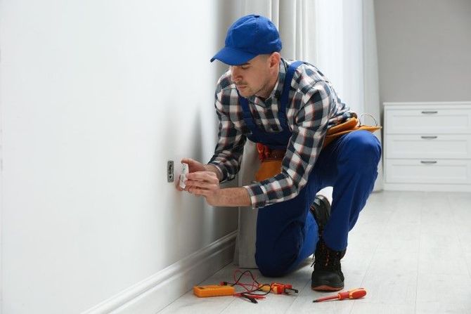 Handyman in blue cap kneeling to fix an electrical outlet, with tools on the floor