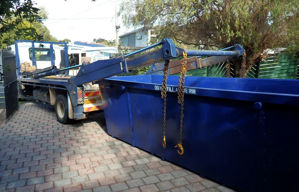 Blue skip bin being lifted by truck's hydraulic arm on a brick driveway. — Select Site Solutions in Kangaroo Valley, NSW
