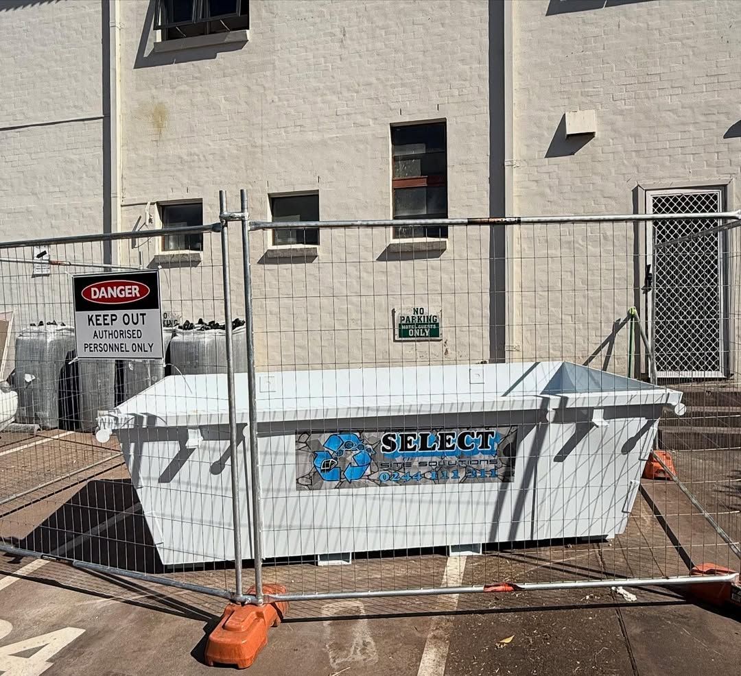 White dumpster behind a metal fence, next to a building with windows. 