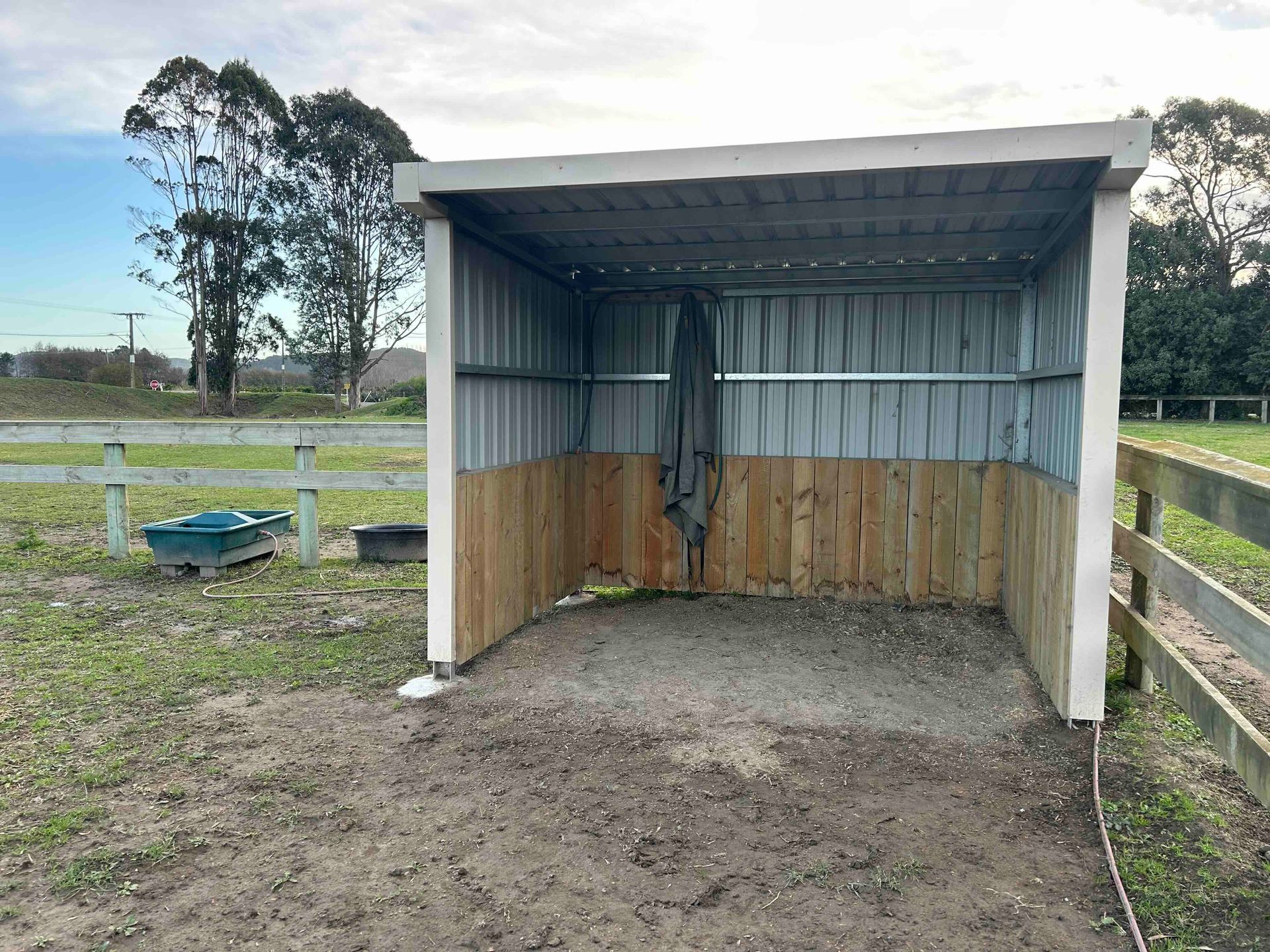 Open-sided shelter made of metal and wood in a rural setting with trees and fencing.