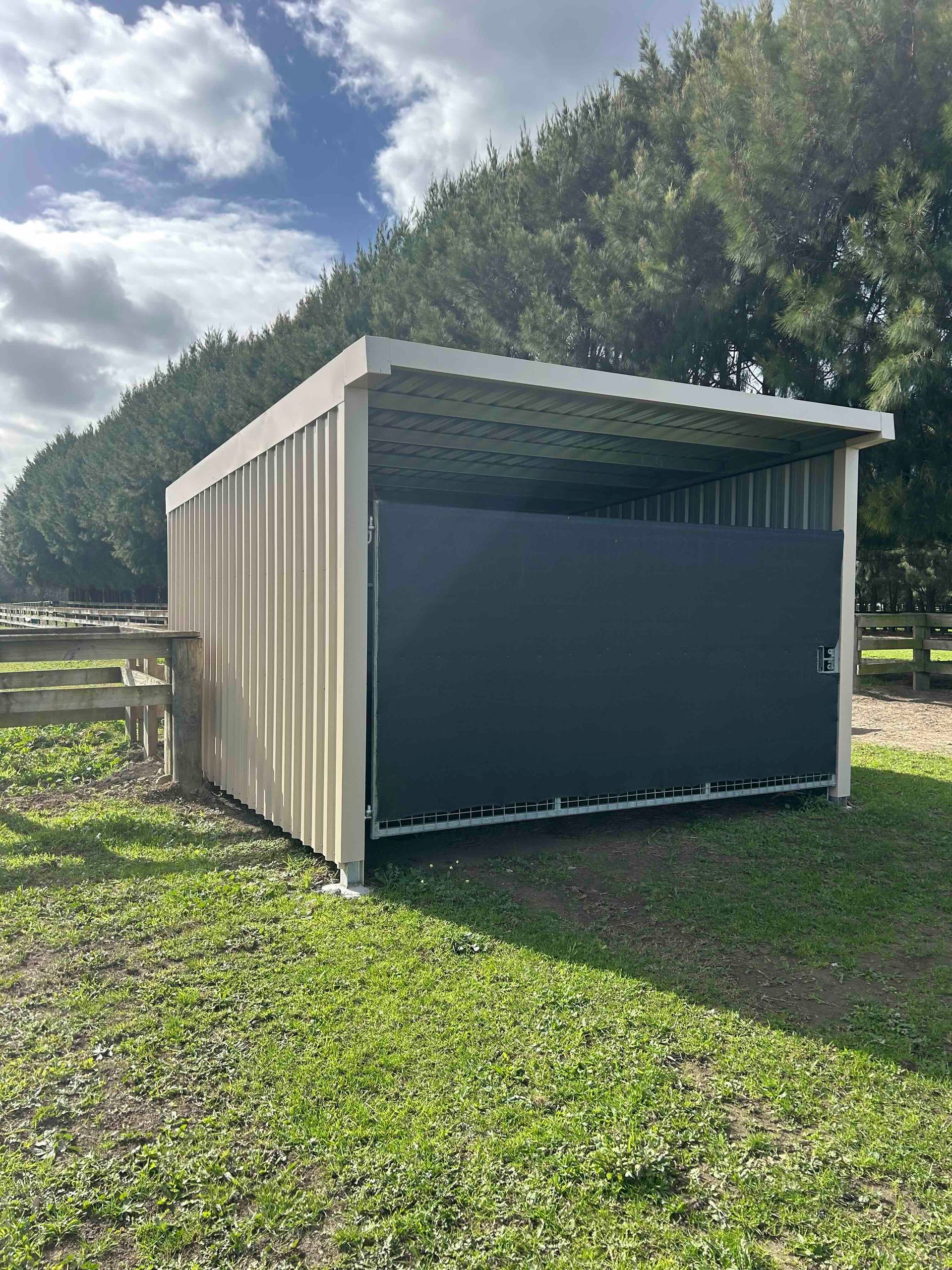 Tan metal shed with black front panel, open to a grassy area, trees in the background.