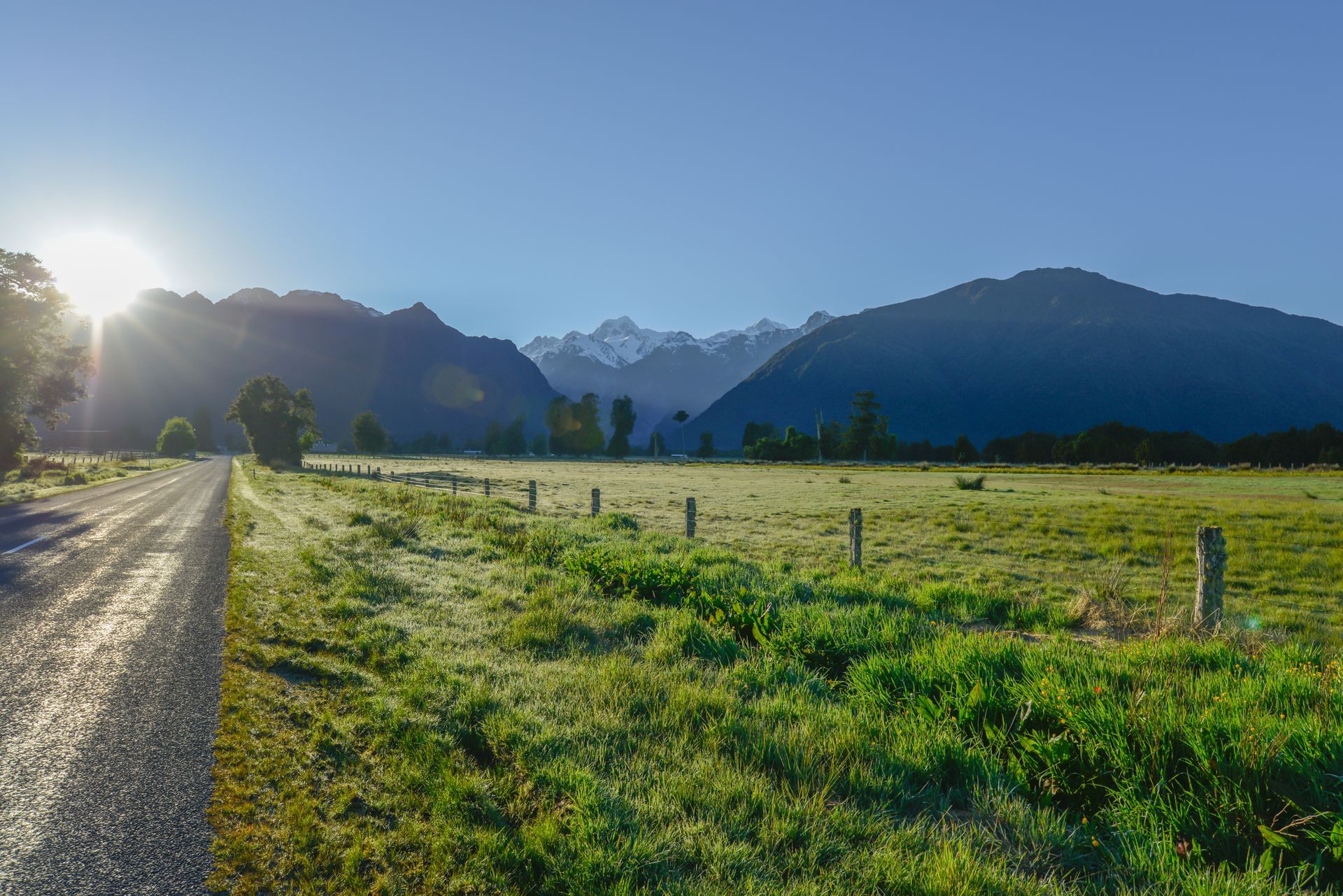 Gravel road through a green field with a fence. Mountains and sun in background.