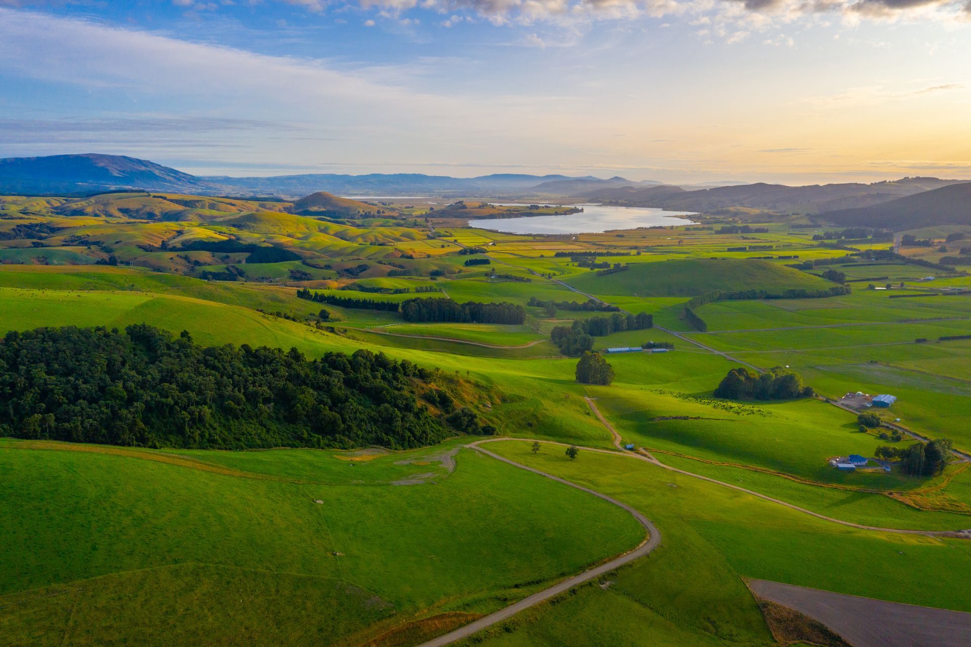 Rolling green hills, a distant lake, and a winding road under a blue sky at sunset.
