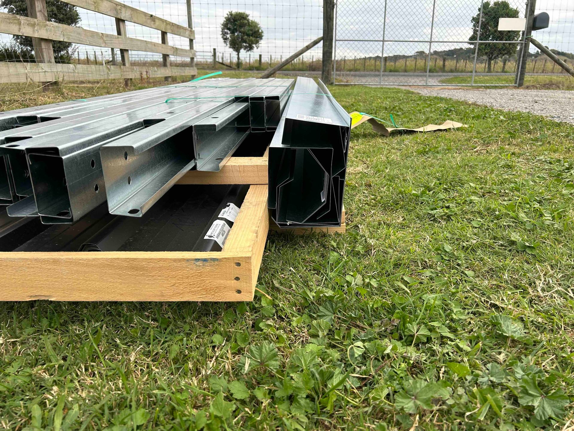 Metal framing materials stacked on a wooden base on grass, near a fence.
