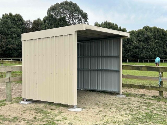 Tan metal shed with open front, in a field with a wooden fence and trees in the background.