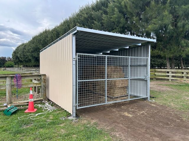 Hay shed with metal roof and wire gate, beige walls. Hay bales visible inside. Outdoors, on grass.