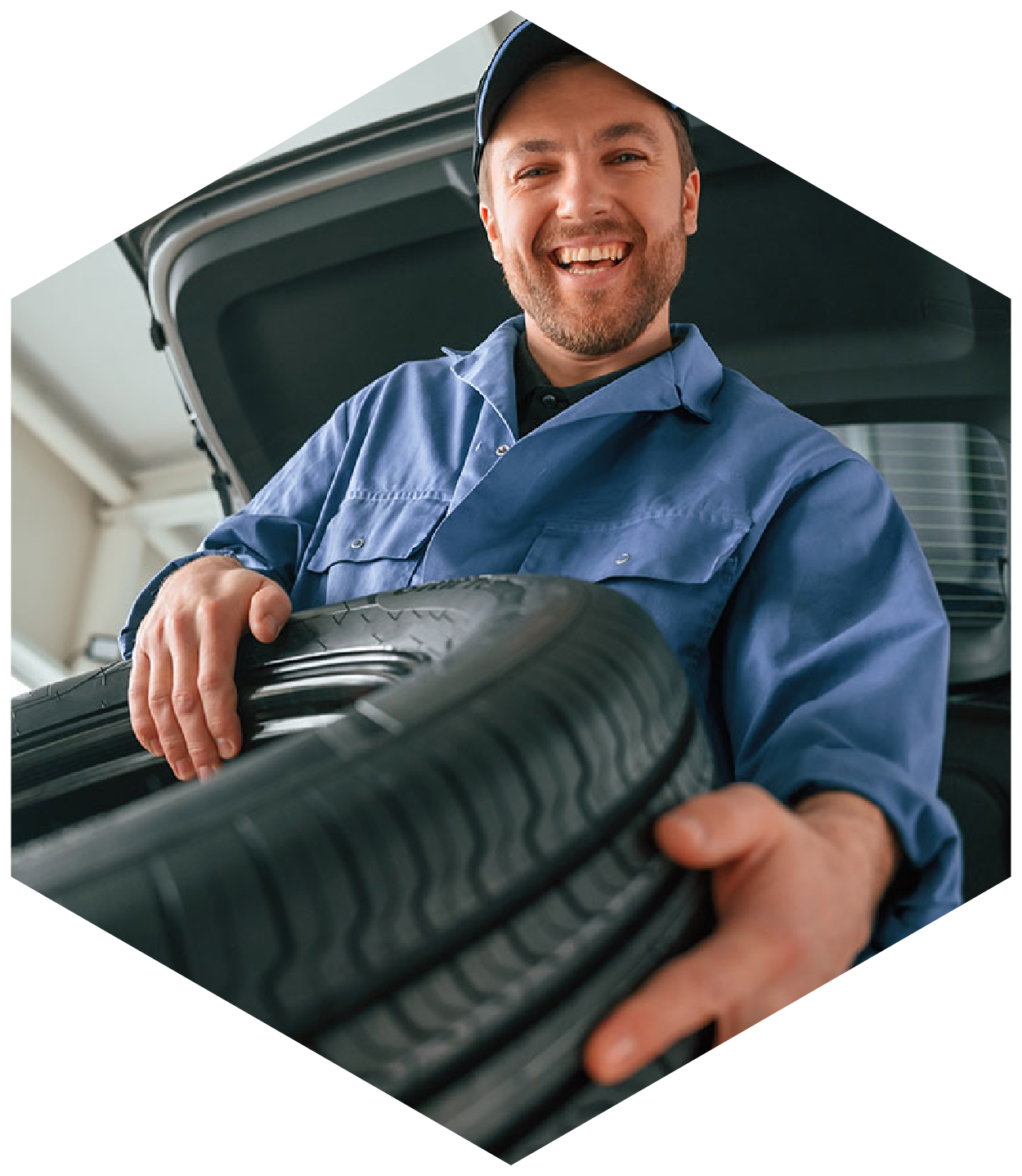 Mechanic holding a tire; smiling in front of an open car trunk. Blue work uniform, indoor setting.
