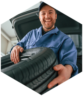 Mechanic holding a tire; smiling in front of an open car trunk. Blue work uniform, indoor setting.