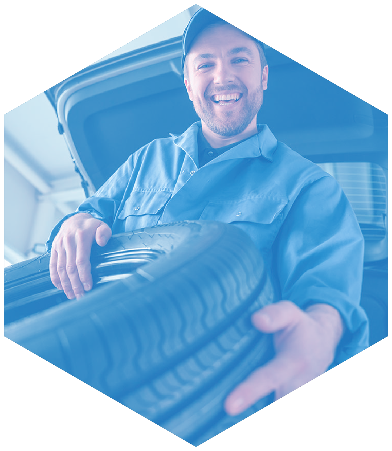 Mechanic smiling, holding a tire; blue uniform, car trunk open, garage setting.