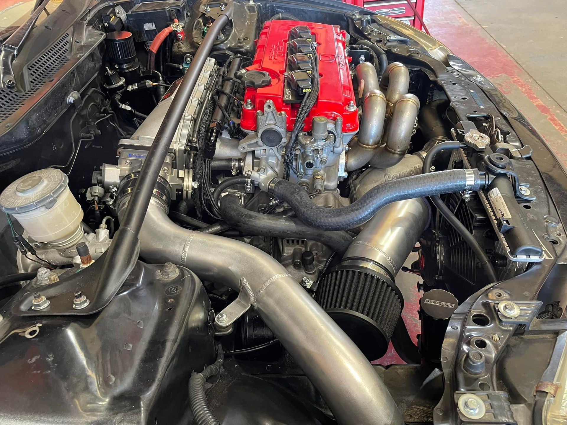 Black car engine bay with red engine components and silver piping, angled view.