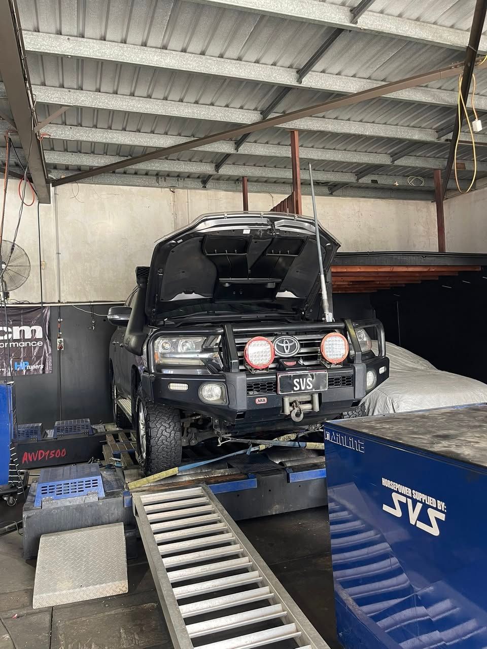 Black SUV with hood open on a dyno in a garage with a ramp.