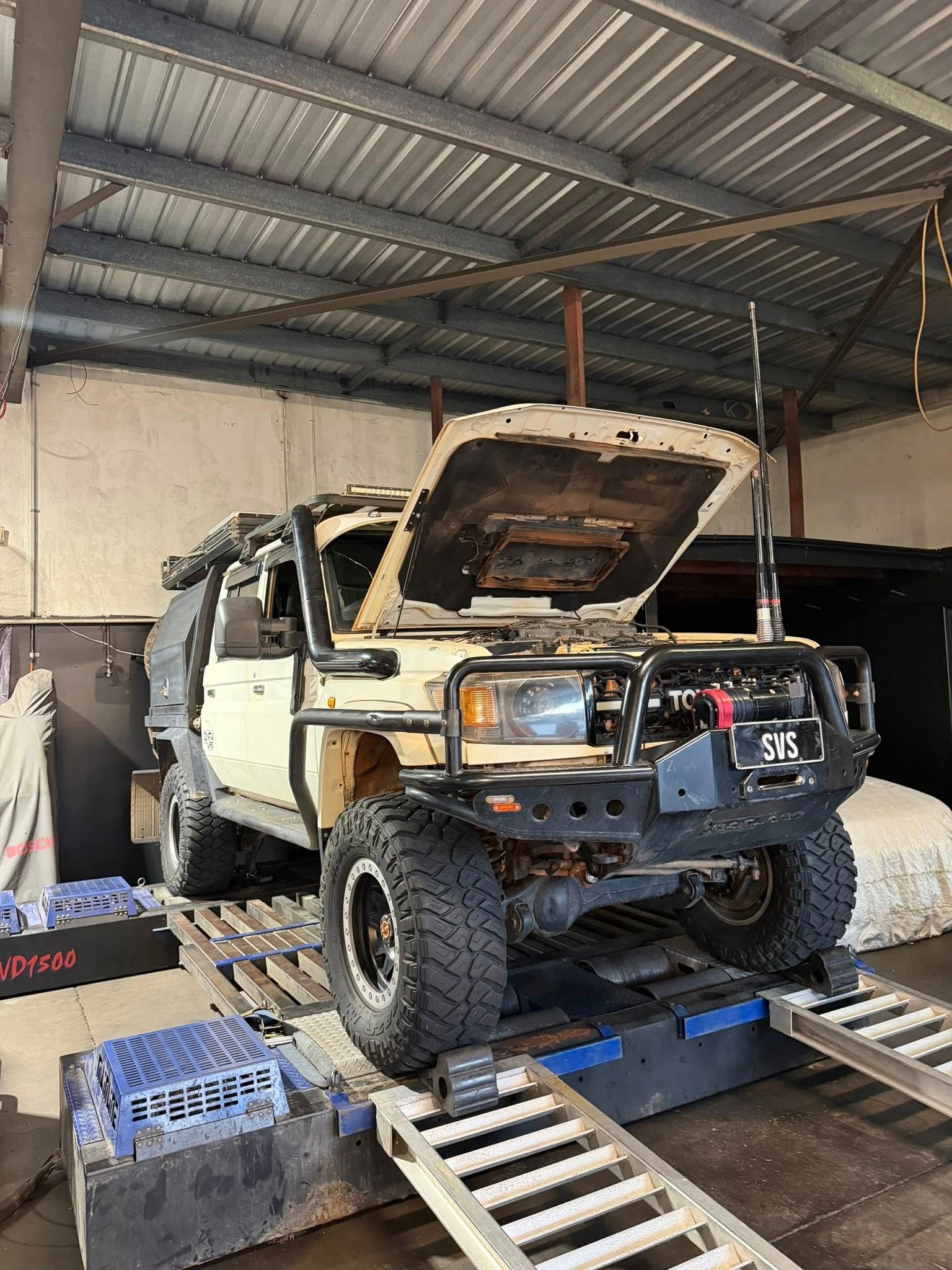 Tan 4x4 vehicle with hood open, on a dynamometer in a garage.