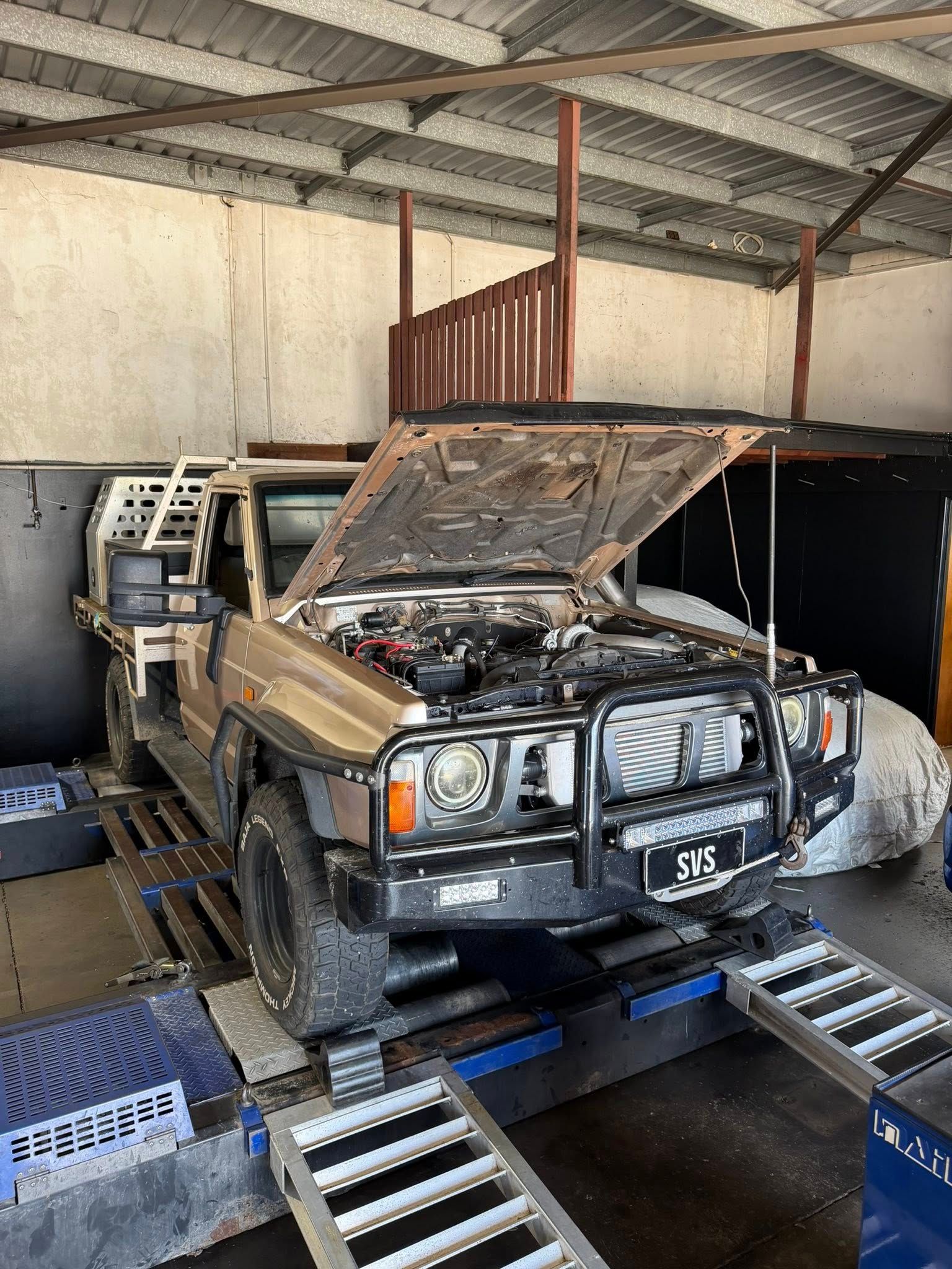 Tan 4x4 vehicle with open hood on a dynamometer in a garage.